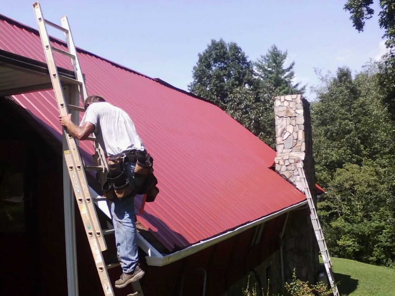 Roofer on a ladder beside a red metal roof and stone chimney — Soffit & Fascia Repair by Roofing Brothers in Brooker
